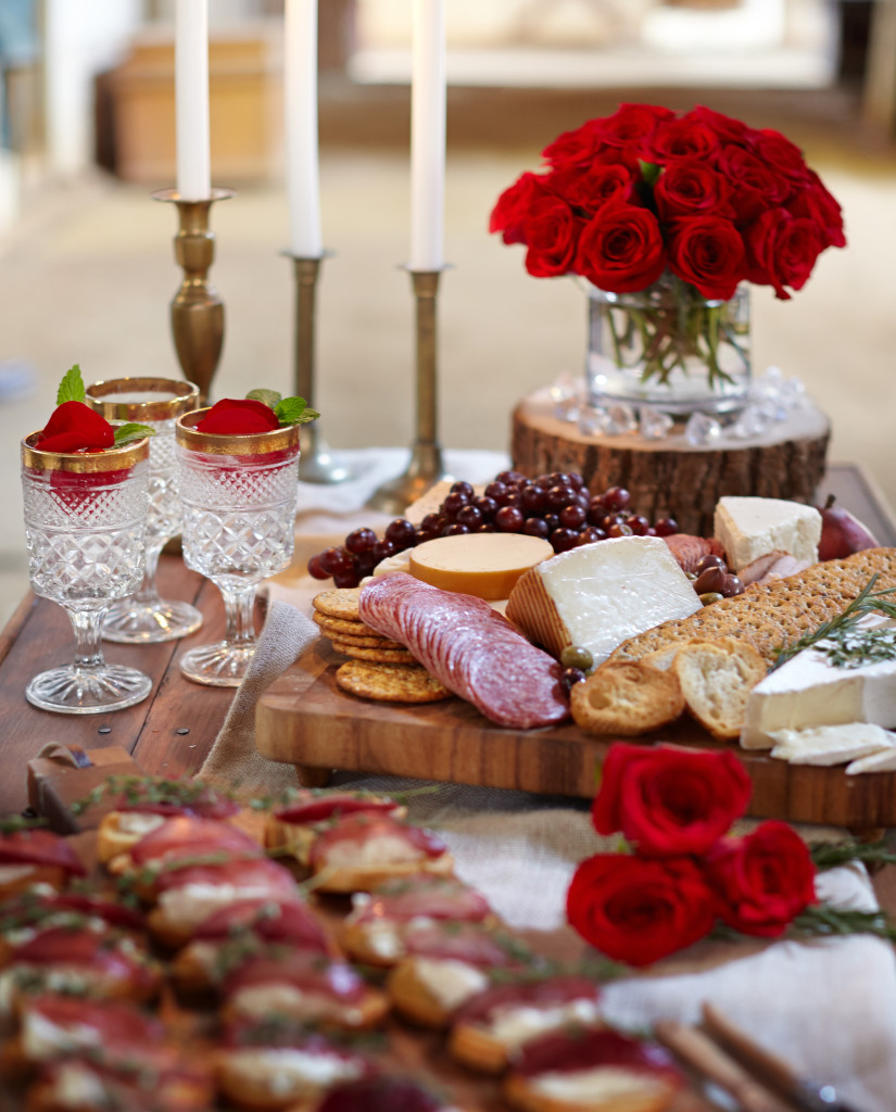 Table setting with wooden cutting board and accents, crystal glassware, meats and cheeses and crackers, and red roses